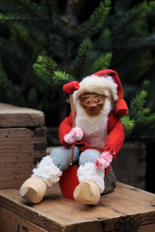 Gammel julenisse med vat skæg , filt tøj og træsko siddende på træstamme. 
Højde: 13 cm.